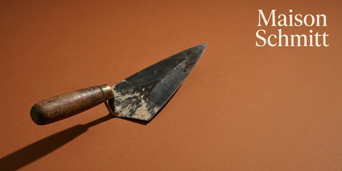Miniature éditoriale du projet Maison Schmitt avec une truelle de maçon en acier patiné au manche de bois huilé posée en diagonale sur un fond ocre terre cuite évoquant le grès rose des Vosges et typographie serif ivoire « Maison Schmitt » sur le tiers droit